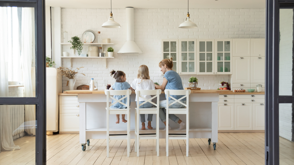 Back view of happy young mother and her little daughters sitting at kithchen table at morning having or cooking breakfast, millennial smiling nanny teaching two girls sisters to prepare food at home